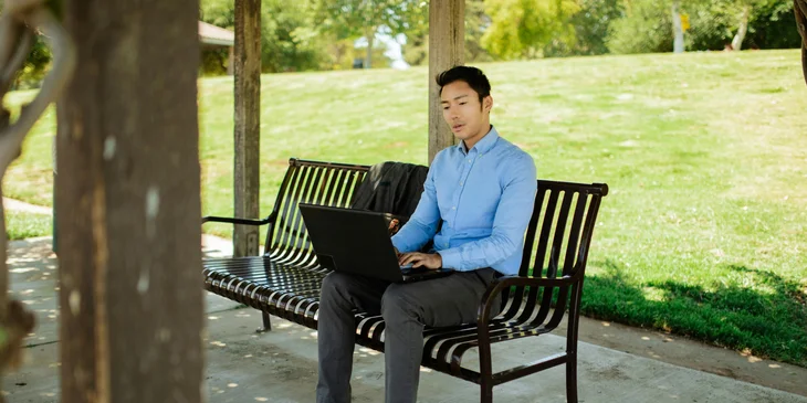 Un hombre trabajando de forma remota en la banca de un parque con su laptop..
