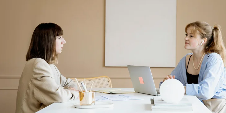 Dos compañeras de trabajo sentadas en una mesa platicando.