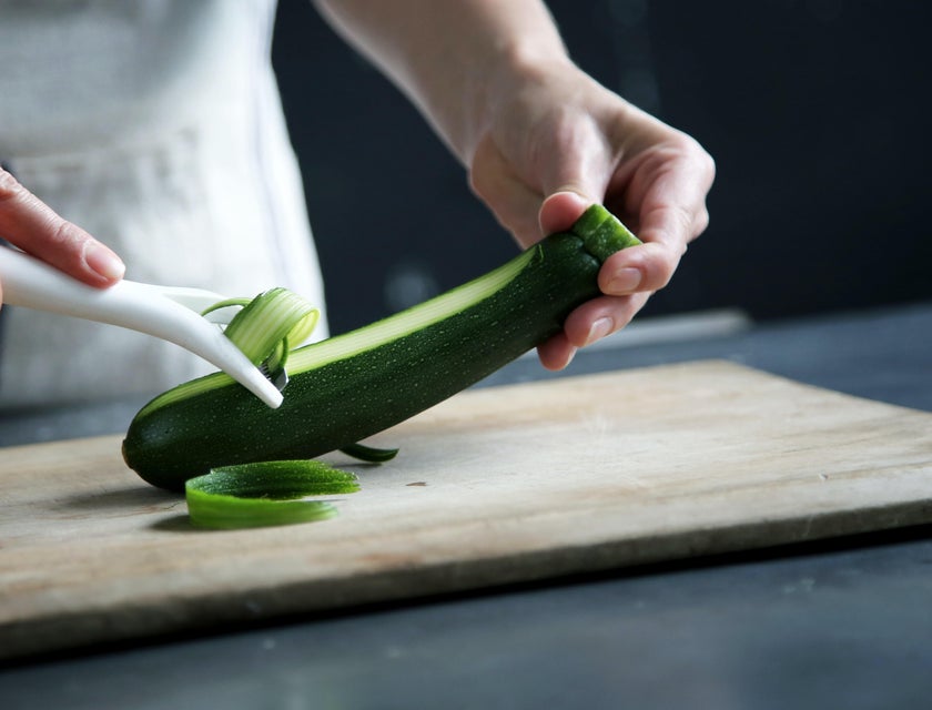 A kitchen staff wearing a white apron peeling a cucumber on a chopping board.