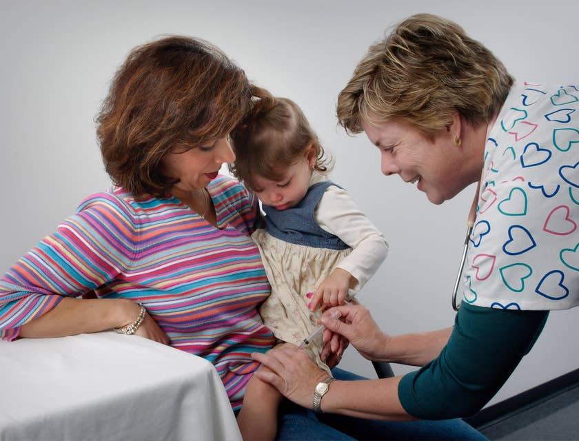 Pediatric medical assistant gives vaccine shot on the leg as the patient's mother comforts her