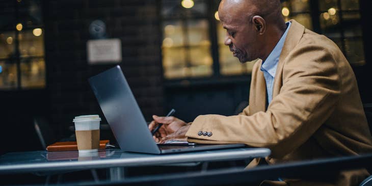 A person writing in a notebook with an open laptop and a cup of coffee.