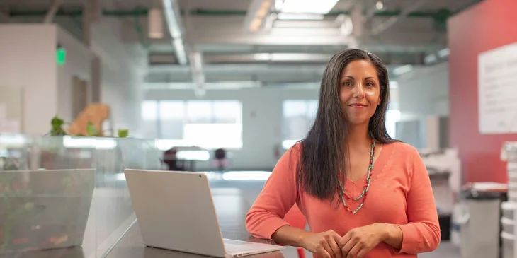 A woman stands next to a desk with a laptop.