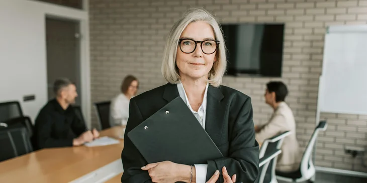 A woman stands holding a clipboard while her coworkers discuss in the background.