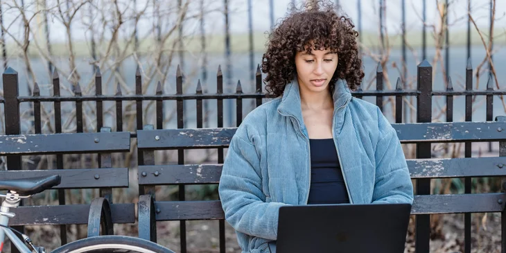 A woman sitting on a bench outdoors, working on her laptop.