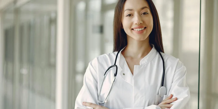 A doctor in medical uniform with stethoscope standing in a clinic corridor.