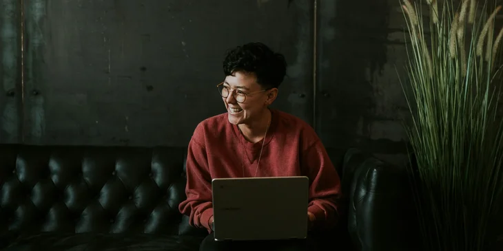 A smiling man sitting on the black sofa with a laptop.