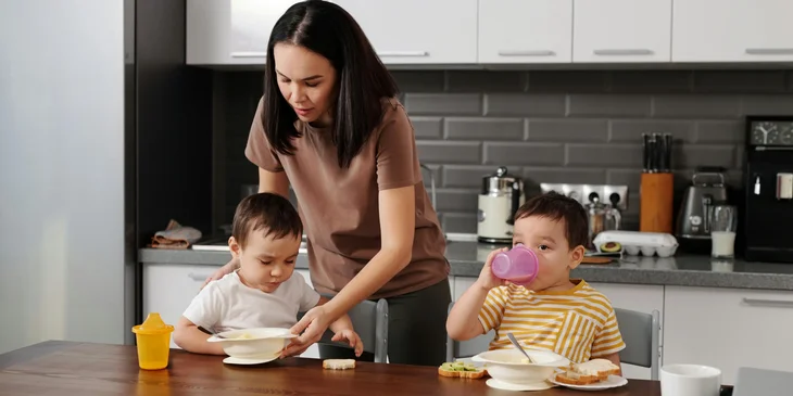 A woman in a brown shirt taking care of the kids in the kitchen.