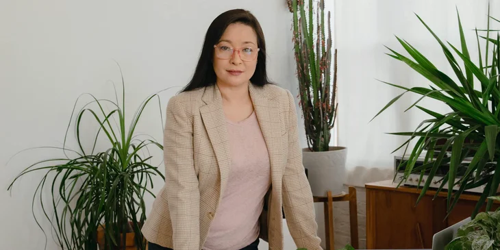 A woman in a beige blazer standing beside a green plant.