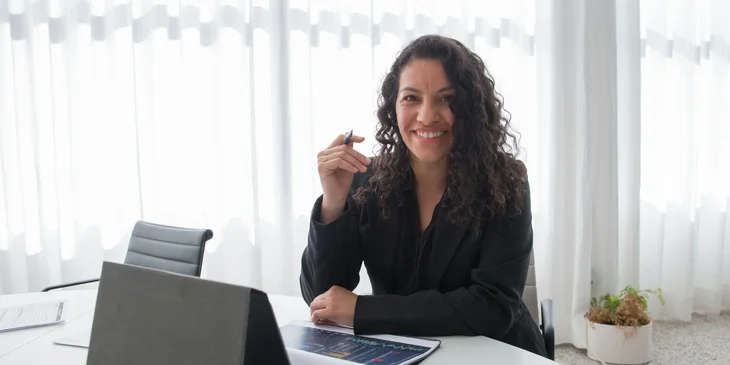 A woman in black blazer sitting by the table smiling.