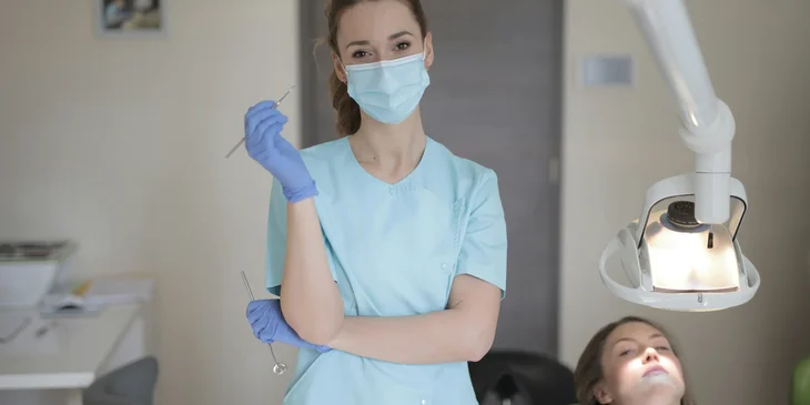 A female dentist wearing a medical mask and gloves standing in a room and looking at the camera.