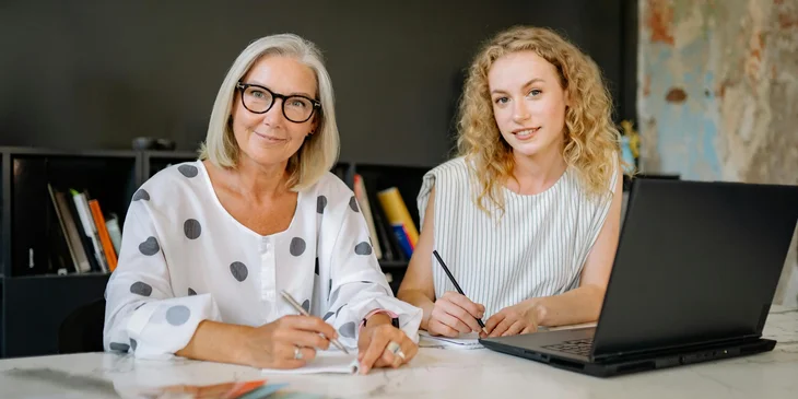 Two smiling women sitting beside each other, each holding a pen in their hand.