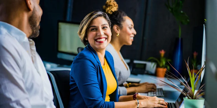 A woman in a blue suit jacket smiling at a coworkwer while working.