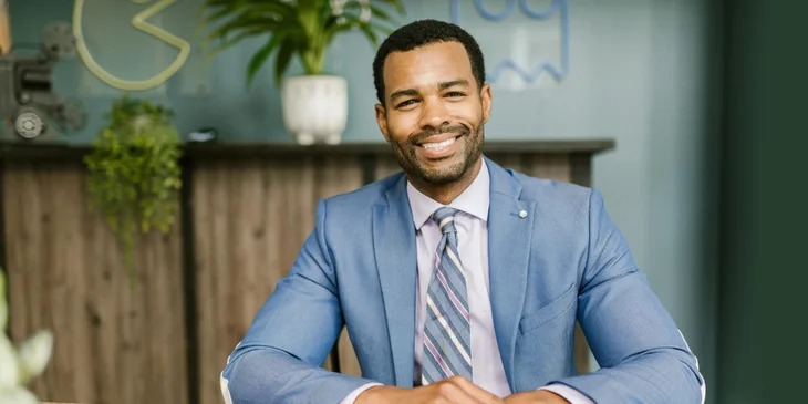 A smiling man in a blue suit sitting at his desk.