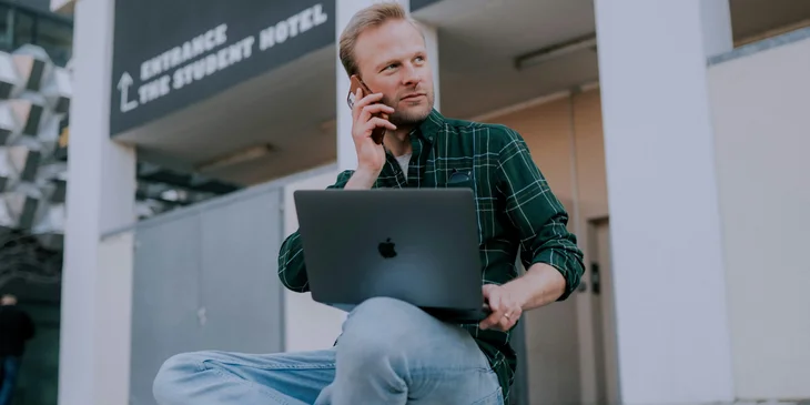 A man in green and black plaid shirt and blue denim jeans sitting.