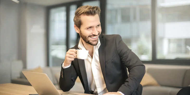 A man holding a white teacup and smiling happily.