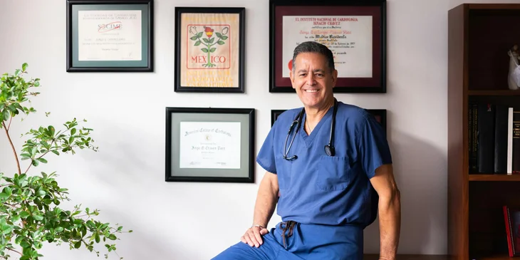 A physician posing in front of a wall filled with framed certificates.