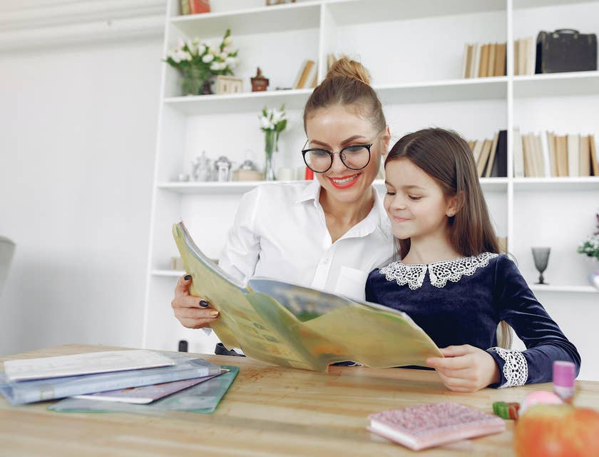 A smiling teacher reading alongside a girl at a desk.