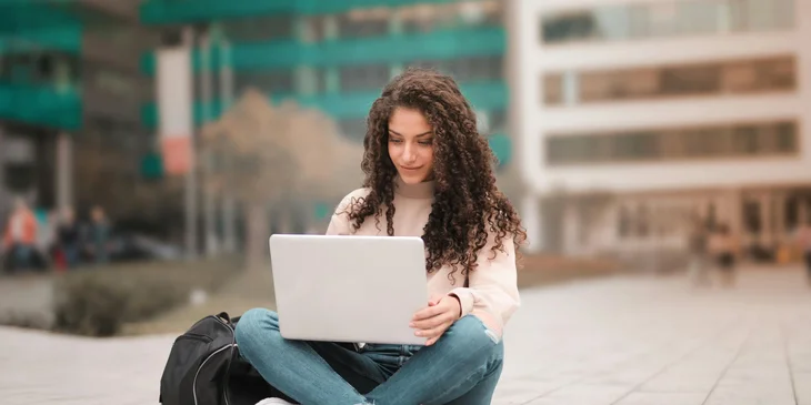 A woman sitting outdoors on the ground, using her laptop.