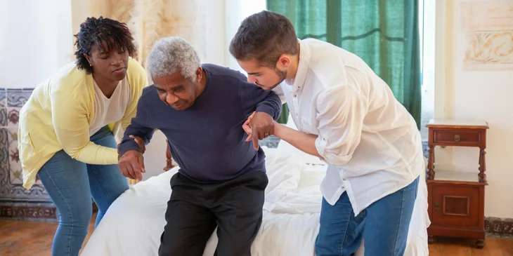 Two home health aides assisting an elderly man in rising from the bed.