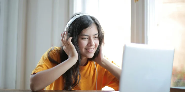 A woman in orange shirt sitting by the table using a laptop.
