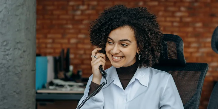 A woman in white long sleeve shirt sitting on a chair while talking on the phone.