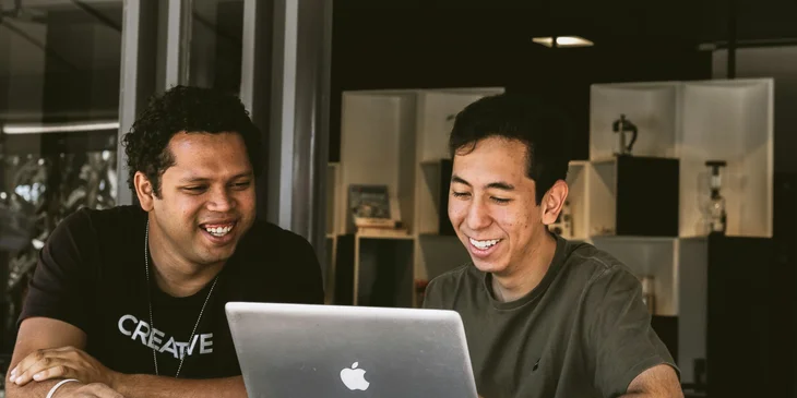 Two Hispanic men happily discussing while browsing on a laptop.