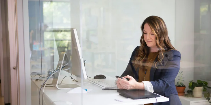 A woman standing by the computer desk and checking her phone.