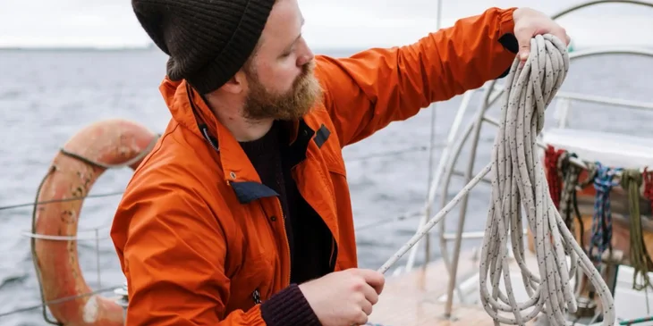 A sailor folding a rope while sitting on a deck.