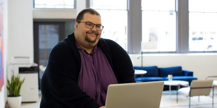 A man sitting on a chair near a table using a laptop.