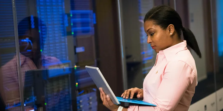 An IT administrator holding a laptop while standing beside a server.