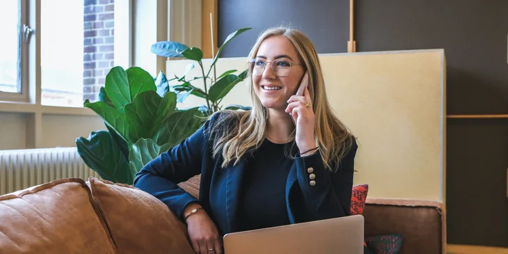 A woman sitting on the couch, speaking on the phone, and holding a laptop.