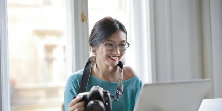 A woman holding a camera and working on a laptop.