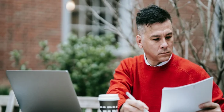 A man in red sweater holding documents and sitting at a table with a laptop on it.
