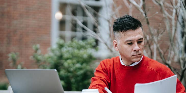 Man in red sweater holding documents and sitting at a table with a laptop on it.