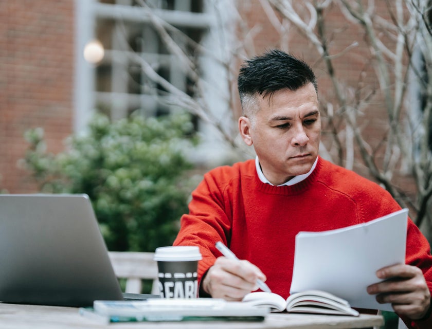 Man in red sweater holding documents and sitting at a table with a laptop on it.
