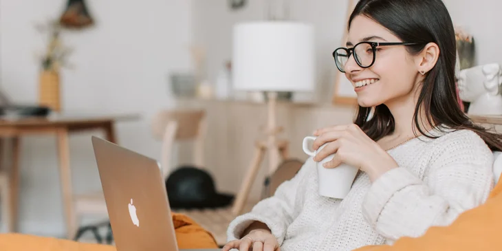 A woman seated on a sofa, drinking coffee while working on her laptop.