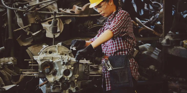 A woman wears a yellow hard hat while holding a vehicle part.