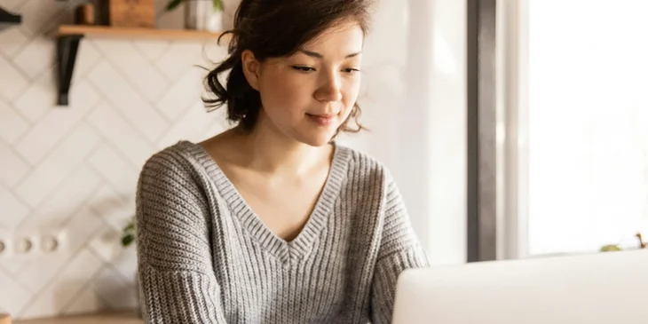 A smiling young woman using a laptop in a kitchen before breakfast.