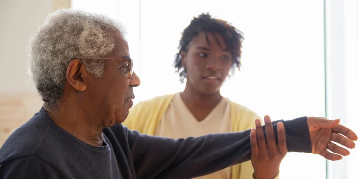 A patient sitter helps the elderly man with stretching exercises.