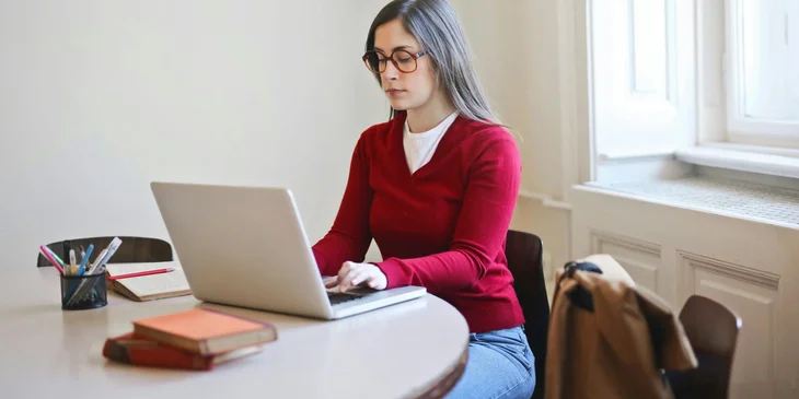 A woman in red long sleeve shirt sitting on a chair while using a laptop.