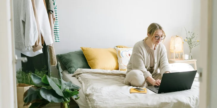 A woman working remotely while sitting on her bed.