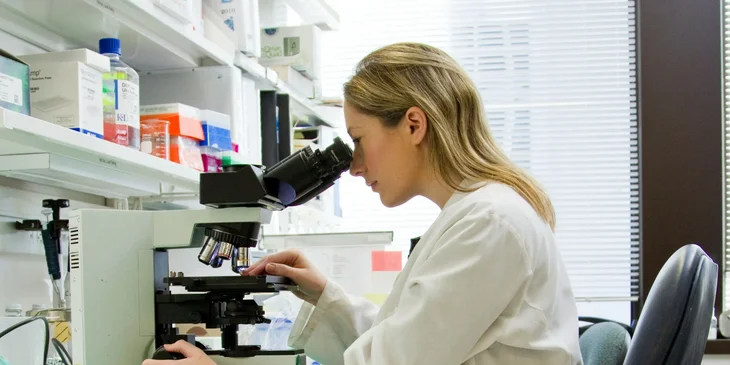 A woman in a white robe looking into a microscope.