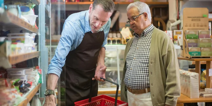 A senior sales representative assisting an elderly man with his vegetable selection at the grocery store.