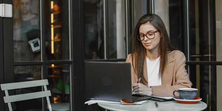 A woman wearing glasses sitting in a coffee shop using her laptop and taking notes.