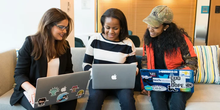 Three women sitting on a sofa with their laptops.