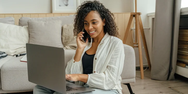 A woman in a white long-sleeved shirt talking on the phone while working on her laptop.