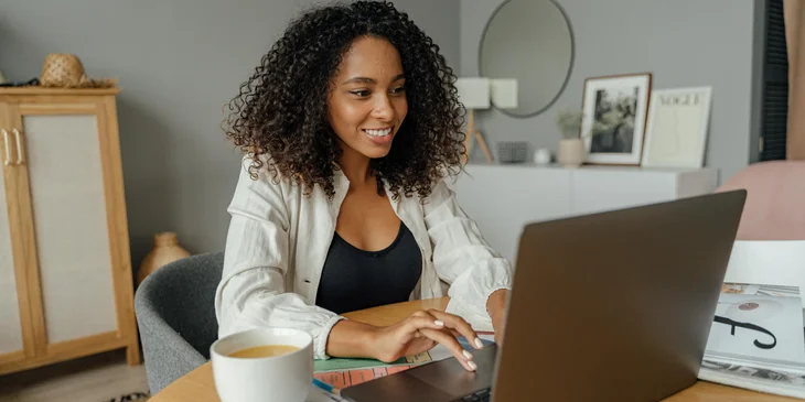 A woman in white blazer using a laptop on a brown wooden table.
