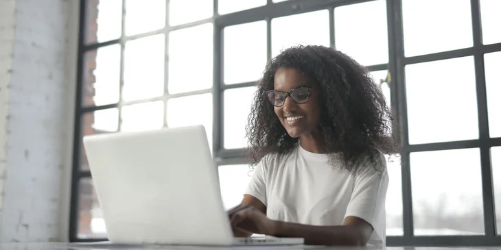 A woman in a white crew neck t-shirt sitting by the window using a laptop.