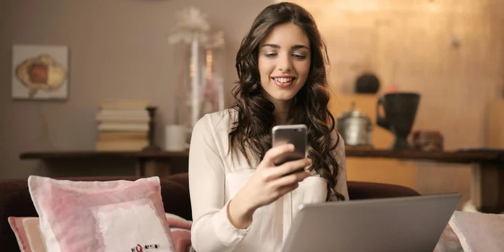 A woman using her mobile phone and laptop while sitting on a sofa.