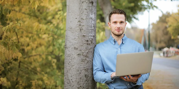 A man standing by the tree holding a laptop.